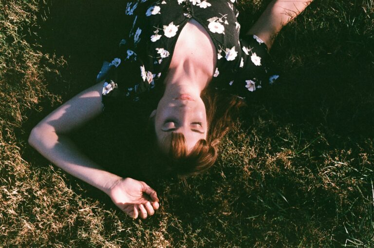 Young woman in a floral dress lying on grass with her eyes closed in warm sunlight