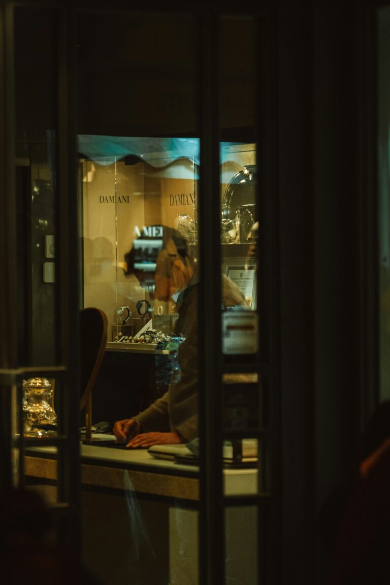 Person sitting alone inside a warmly lit shop window viewed from a dark street at night