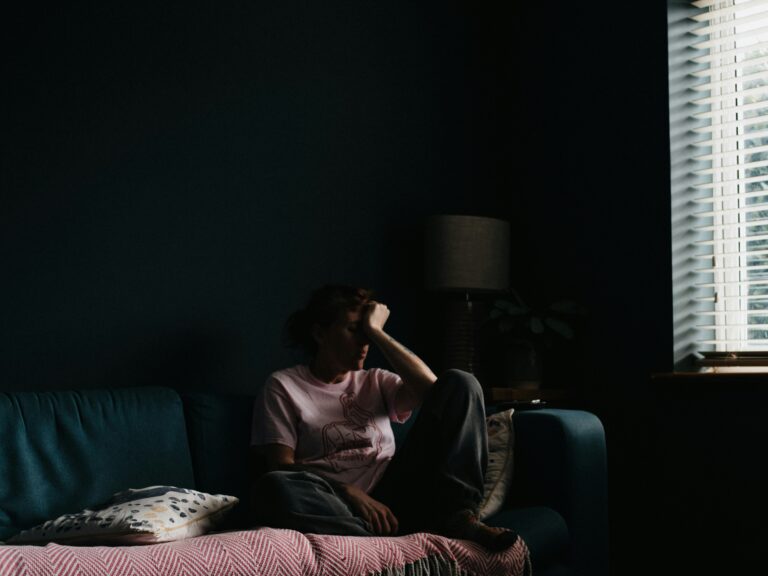 Person sitting alone on a couch in a dimly lit room with light filtering through window blinds