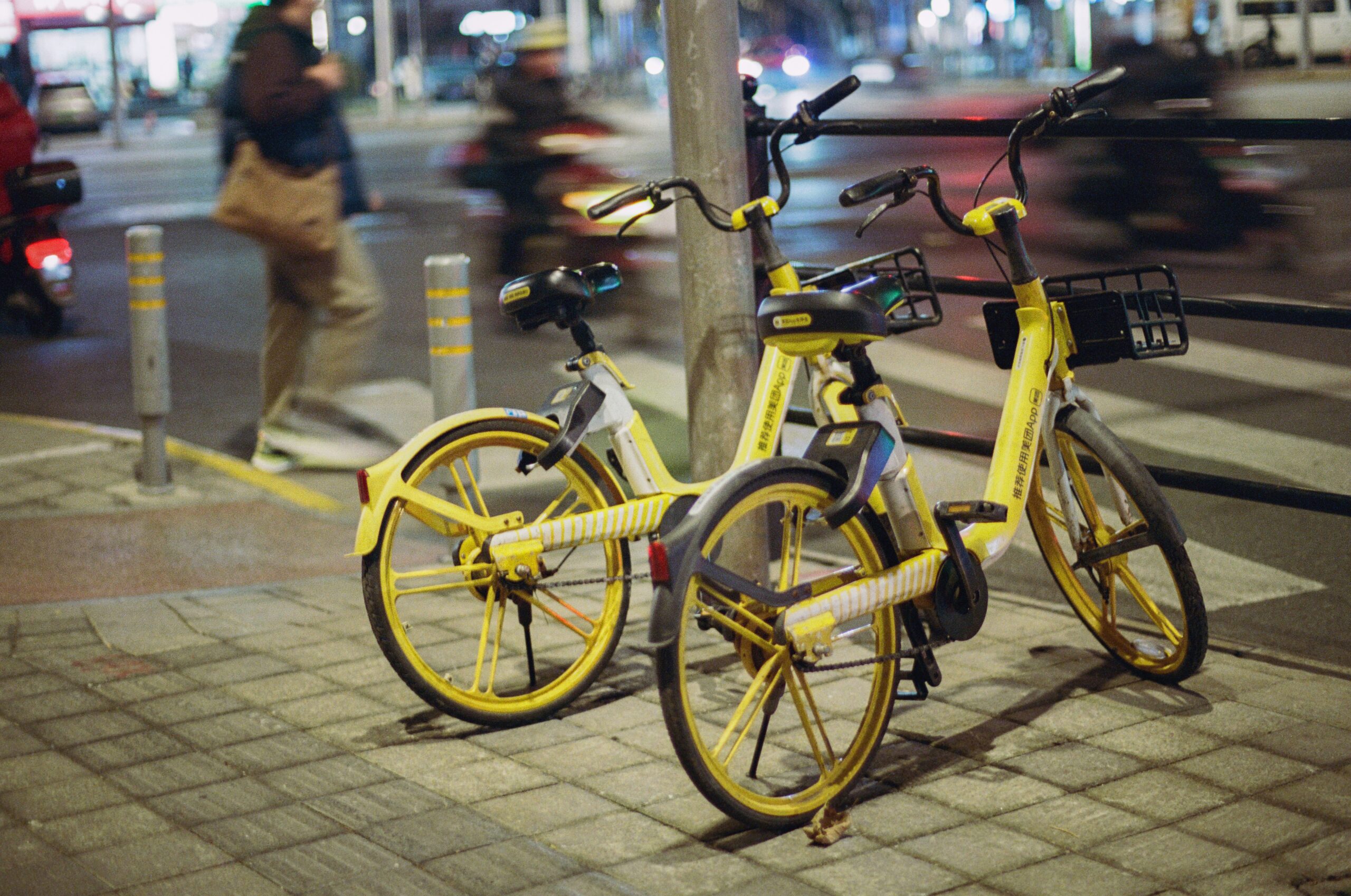 Yellow shared bicycles parked on a city sidewalk at night with blurred pedestrians