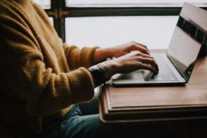 Close-up of a person in a tan sweater typing on a laptop by a window