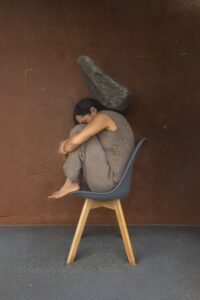 Clay figurine of a person sitting on a wooden chair with a large stone on their back