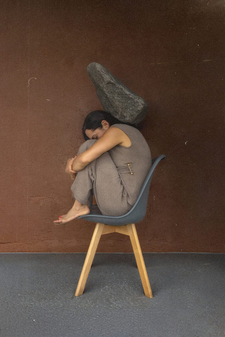 Clay figurine of a person sitting on a wooden chair with a large stone on their back