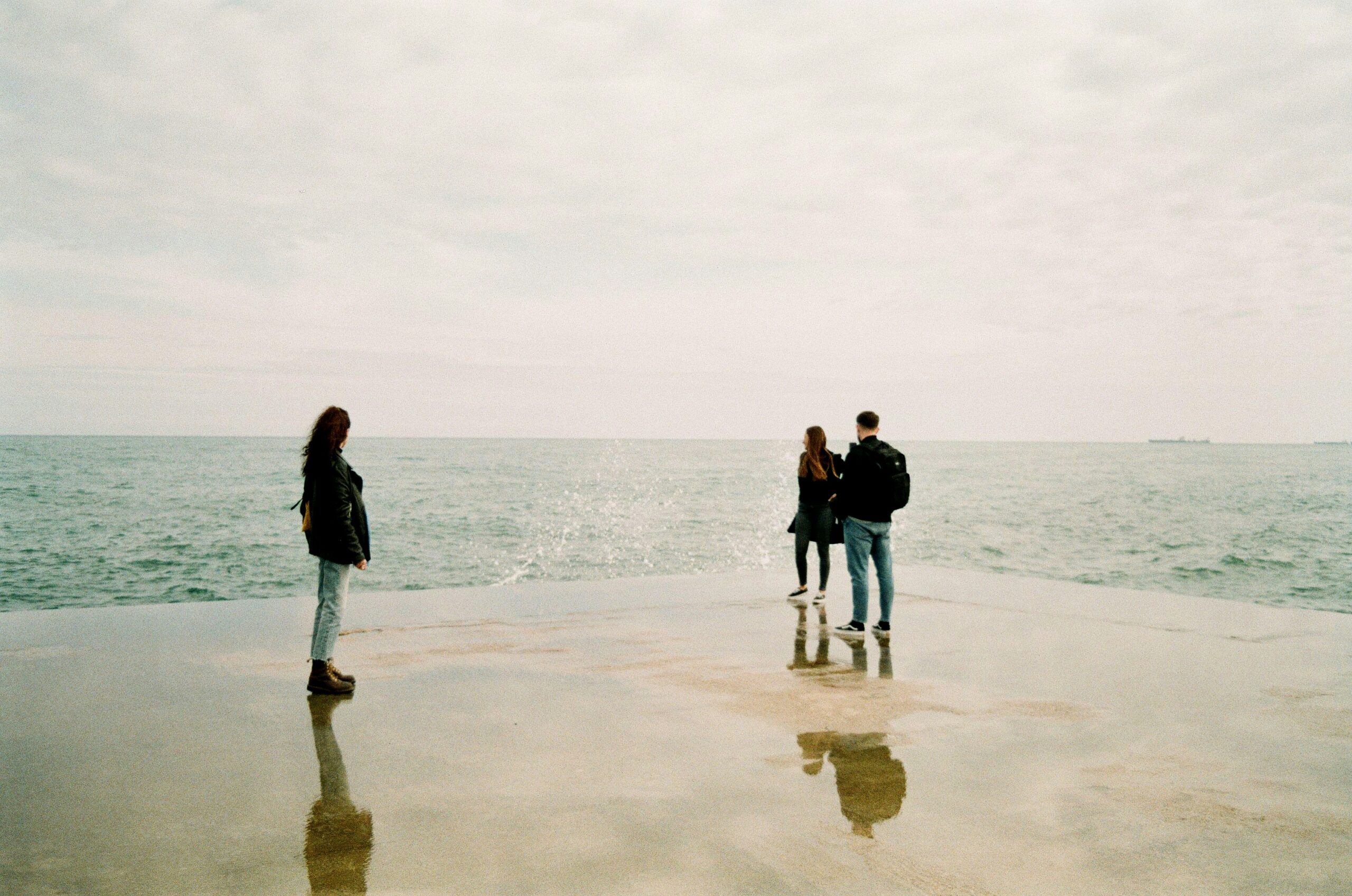 Three young people standing on a wet pier looking out at the ocean on a cloudy day