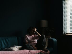 Person sitting alone on a couch in a dimly lit room with light filtering through window blinds
