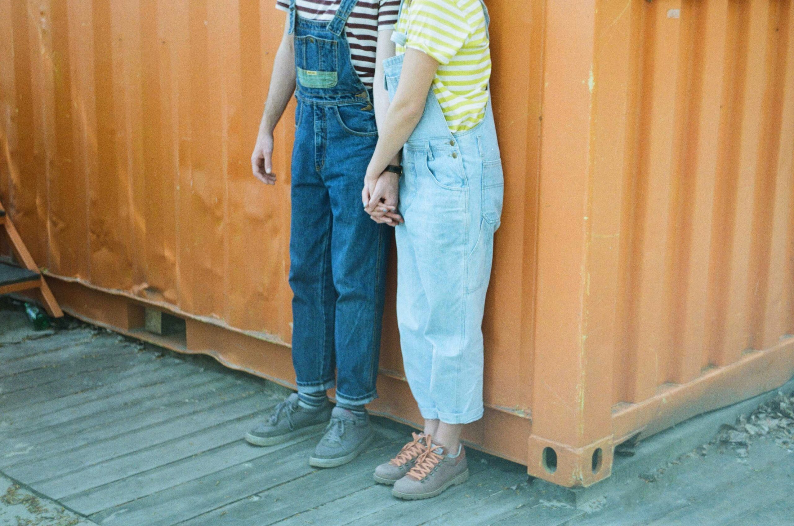 Two people in denim overalls holding hands in front of an orange shipping container