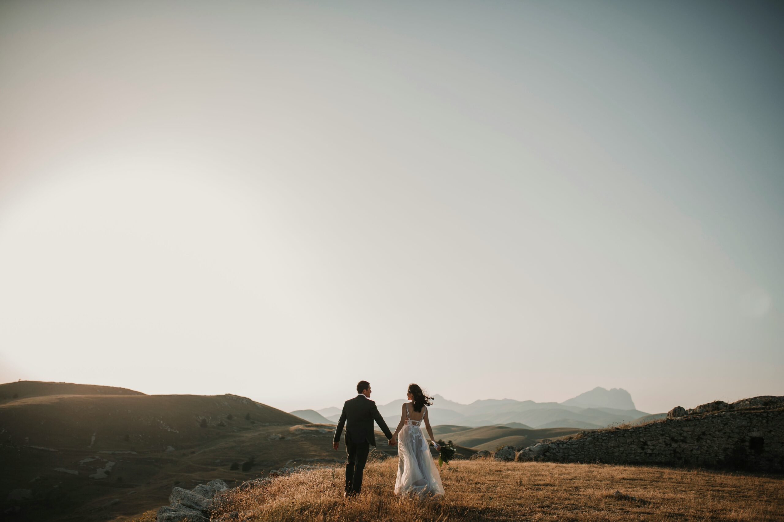 Couple holding hands walking through a golden field at sunset with mountains in the background