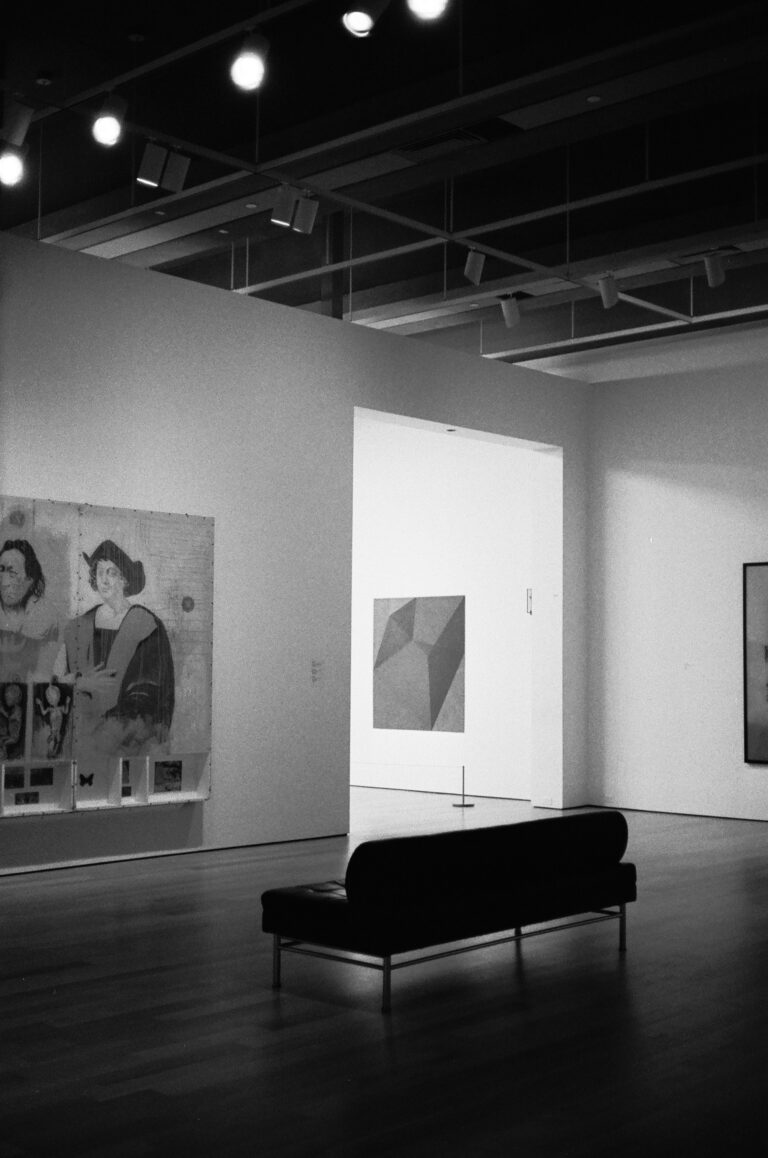 Black and white photograph of an art gallery interior with a bench and paintings on the walls