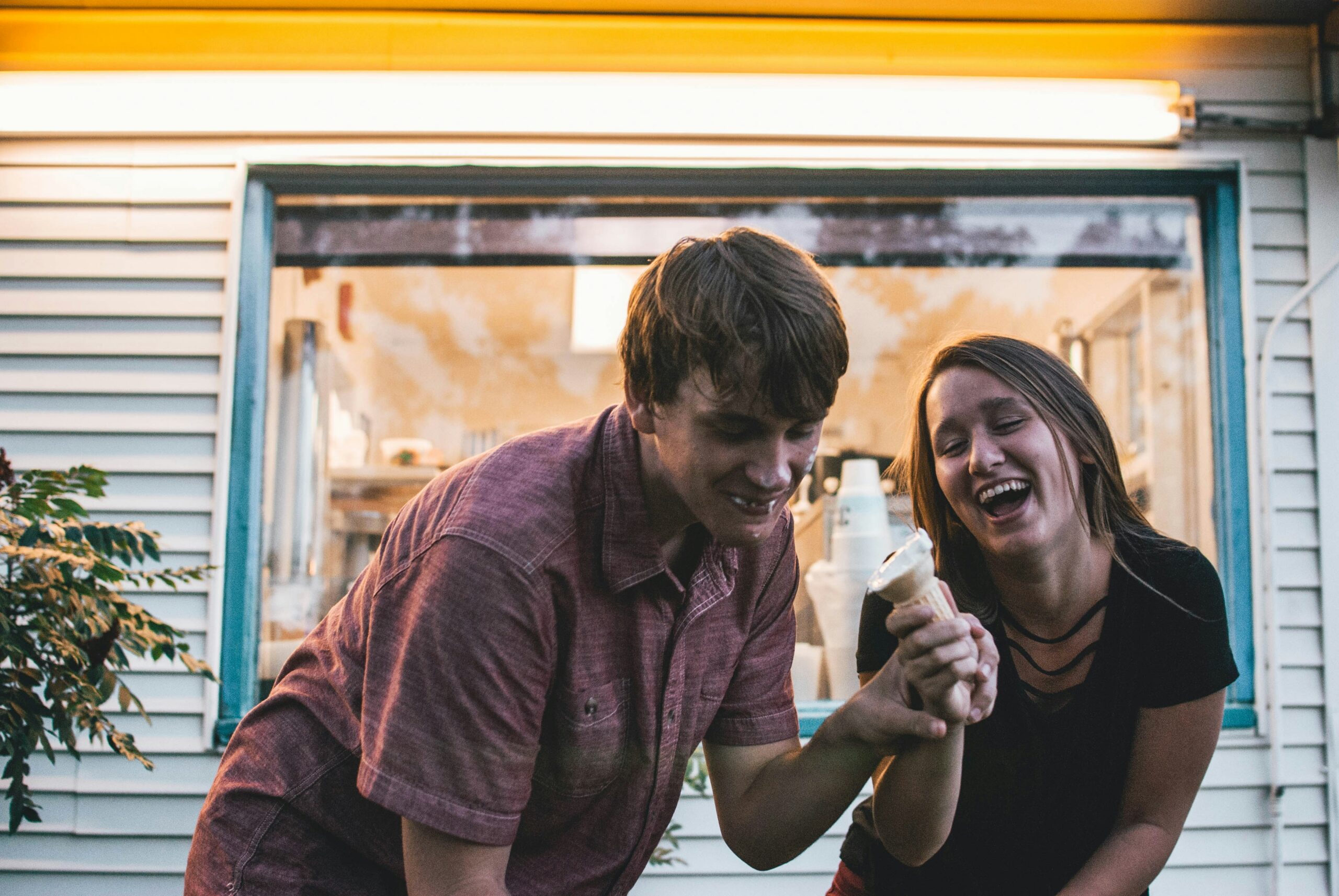 Young Christian couple laughing together while sharing an ice cream cone outside a vintage walk-up window