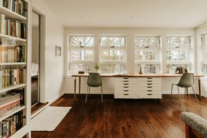 Bright modern home office with floor-to-ceiling windows, sage green chairs, a long wooden desk, and a wall of bookshelves