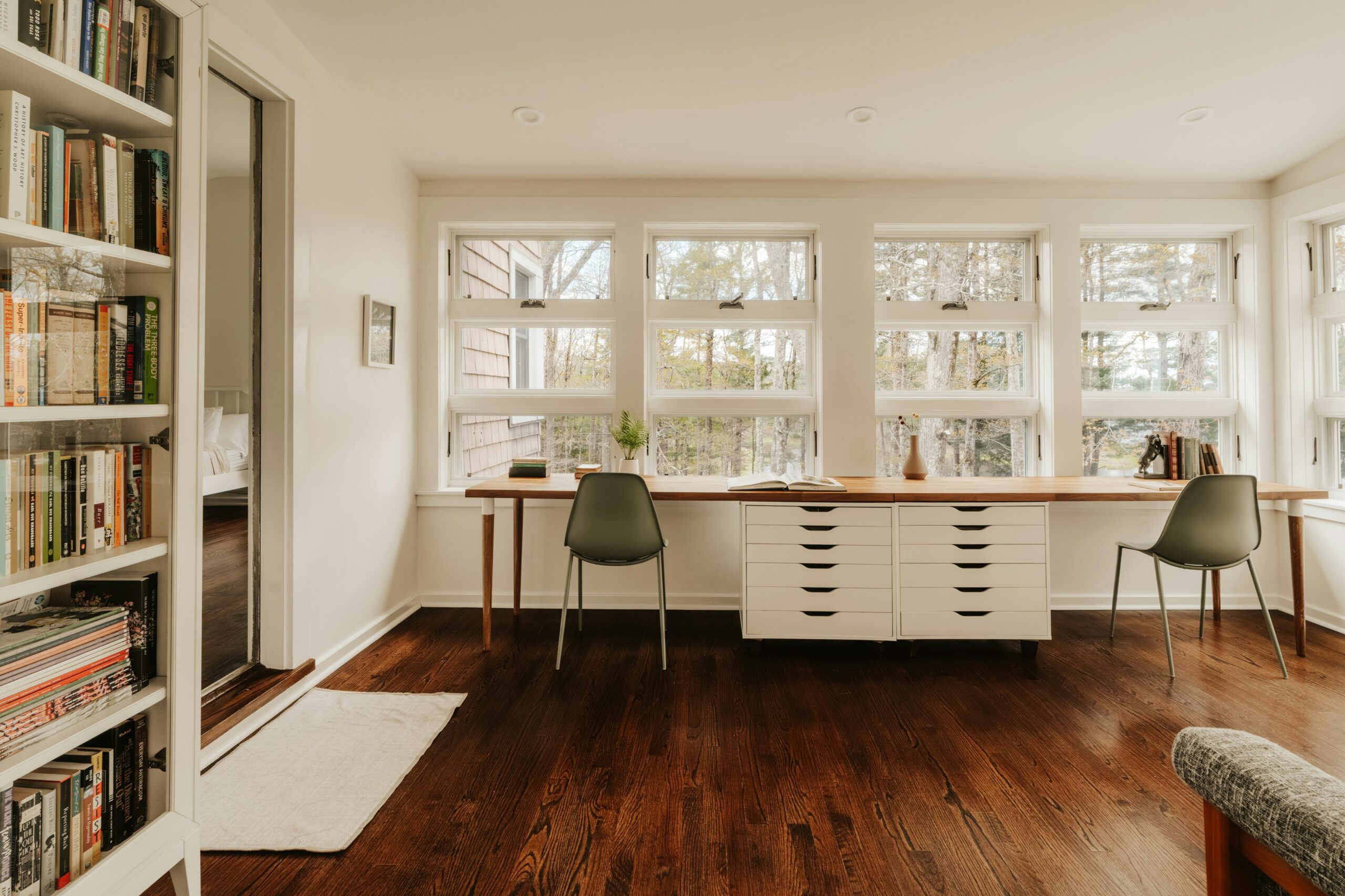 Bright modern home office with floor-to-ceiling windows, sage green chairs, a long wooden desk, and a wall of bookshelves