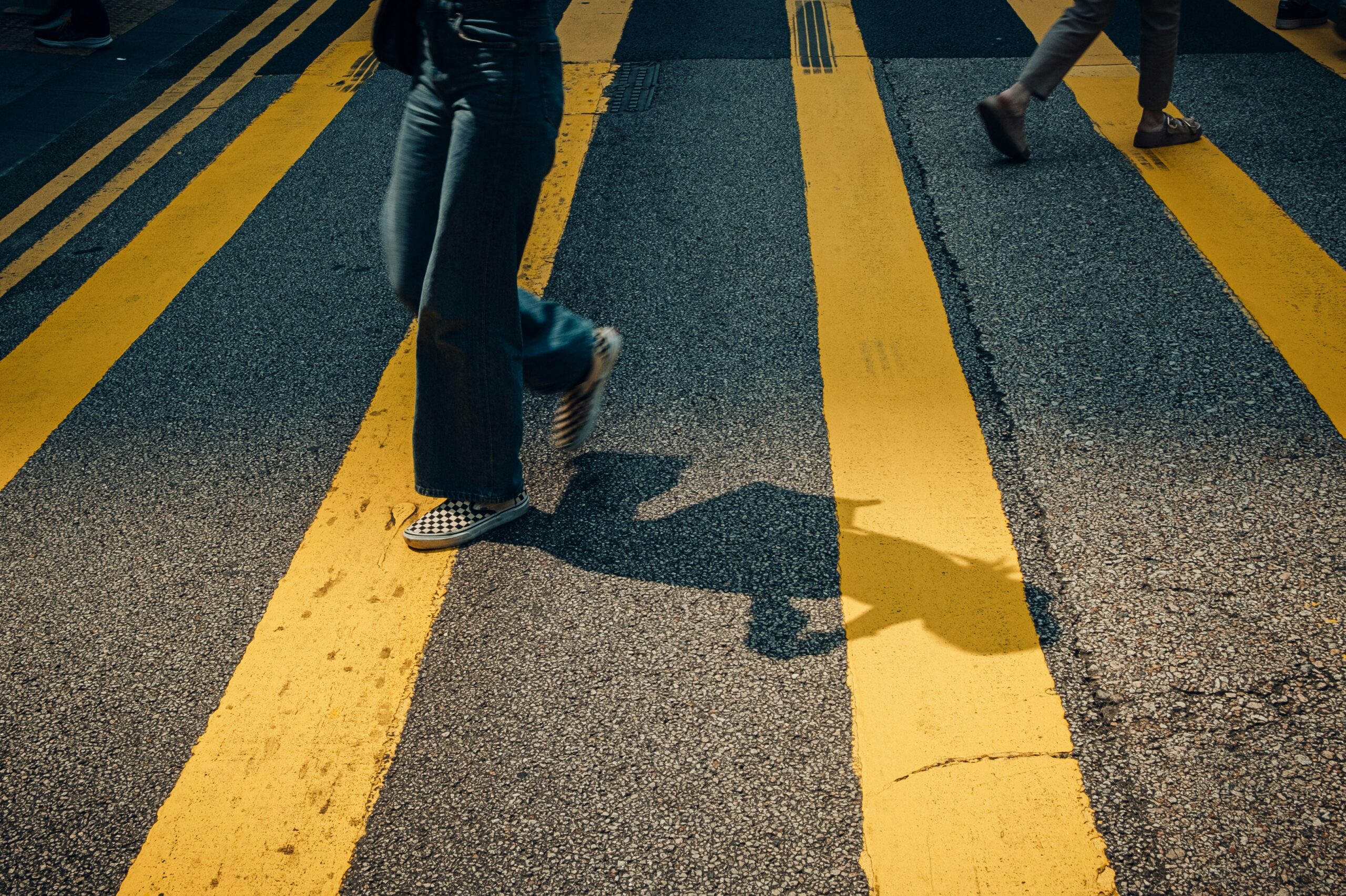 Two people walking across a yellow pedestrian crosswalk with long shadows stretching across the pavement