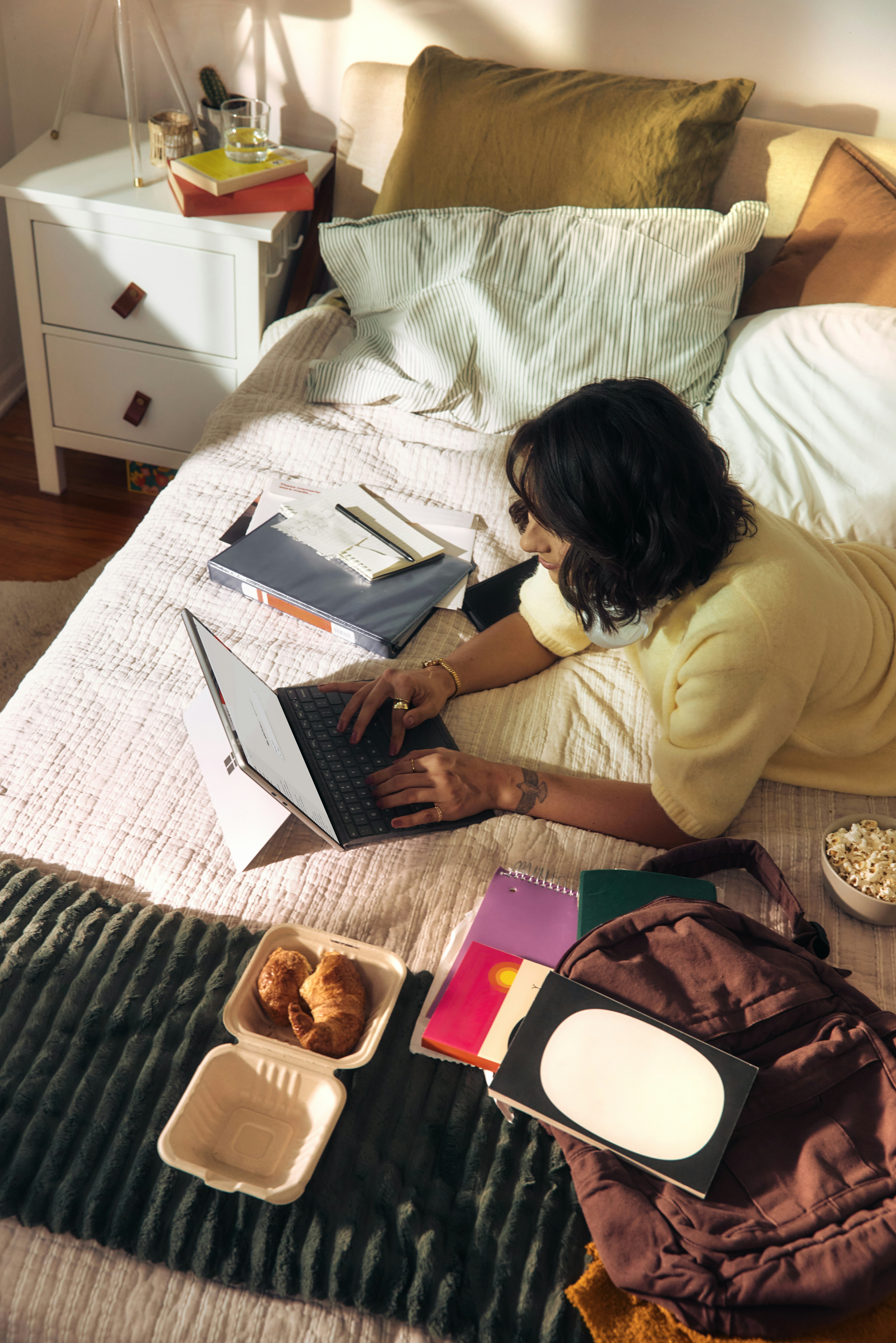 Young woman lying on her bed typing on a laptop surrounded by open notebooks, croissants in a takeout container, and a bowl of popcorn