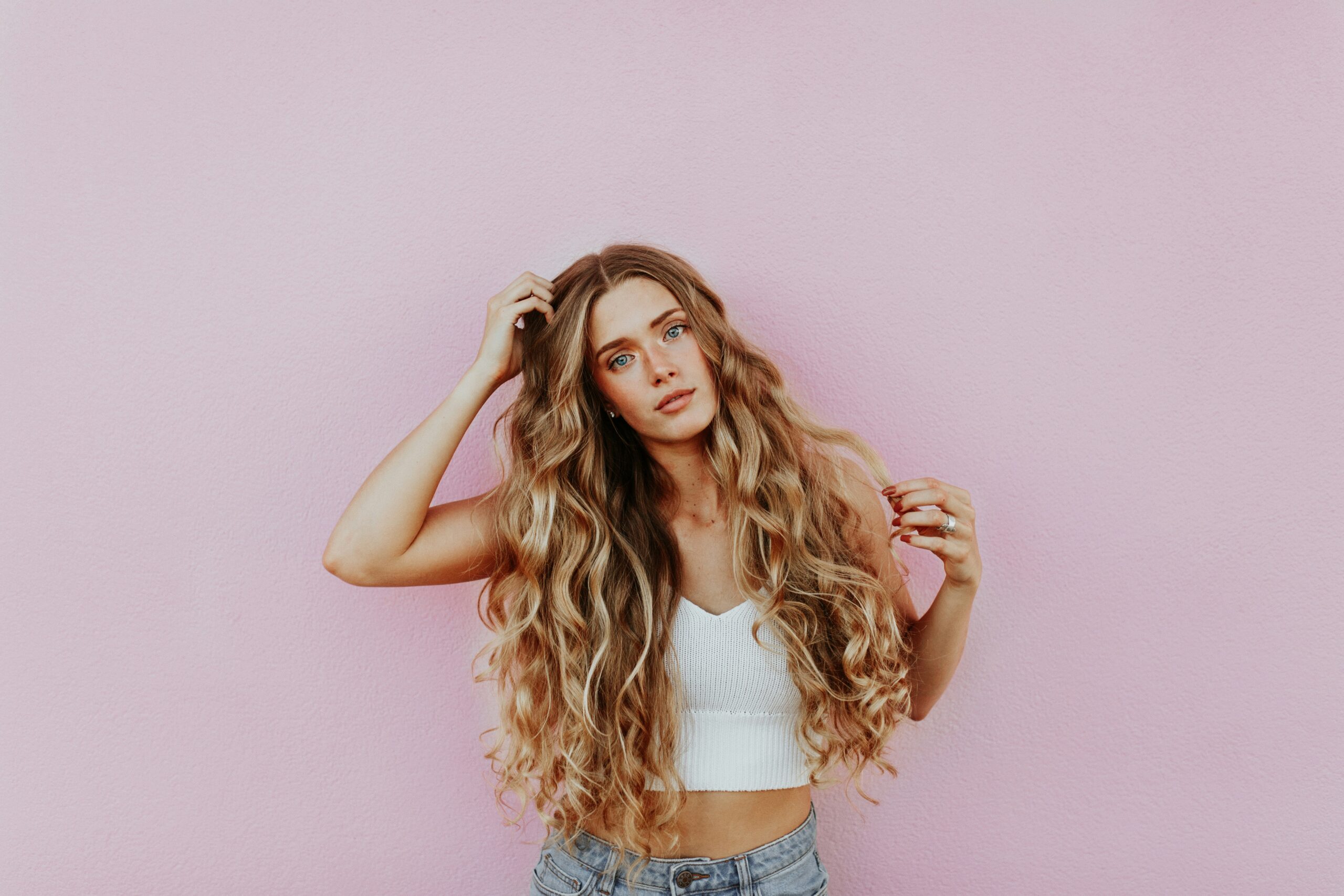 Young woman with long wavy hair in a white top standing against a pink wall, representing freedom from purity culture shame