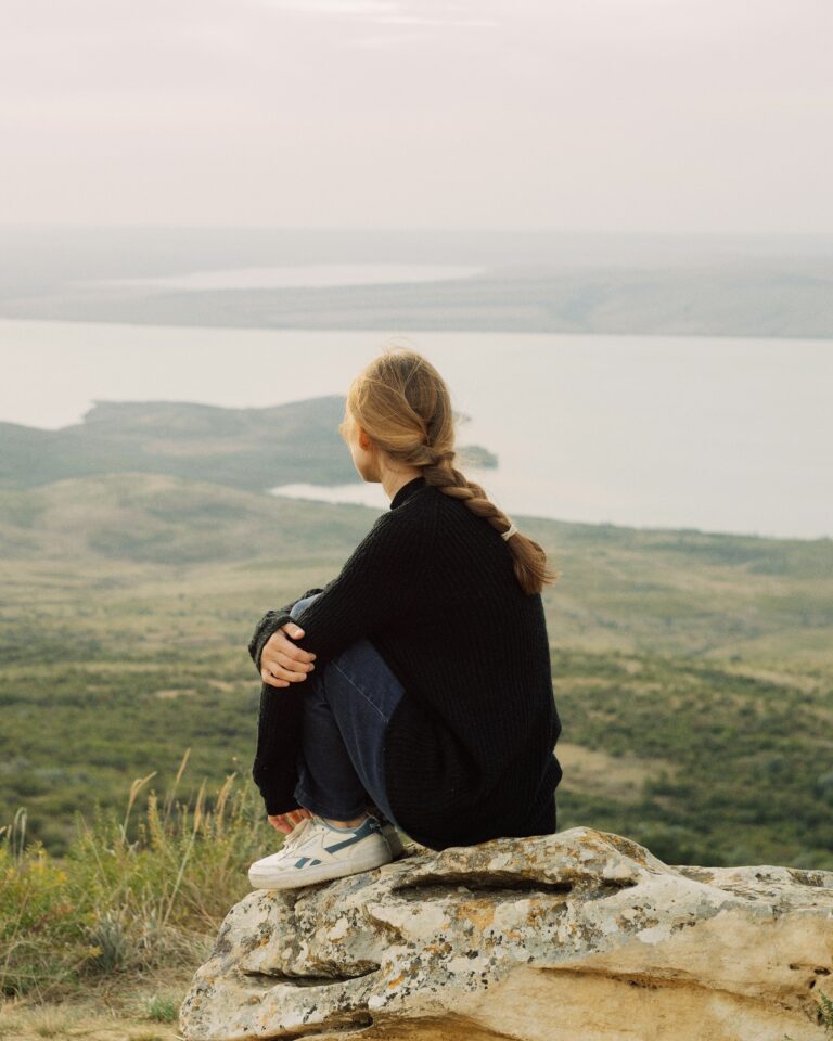 Person with braided hair sitting alone on a rock above a still lake, weighing a decision about a relationship