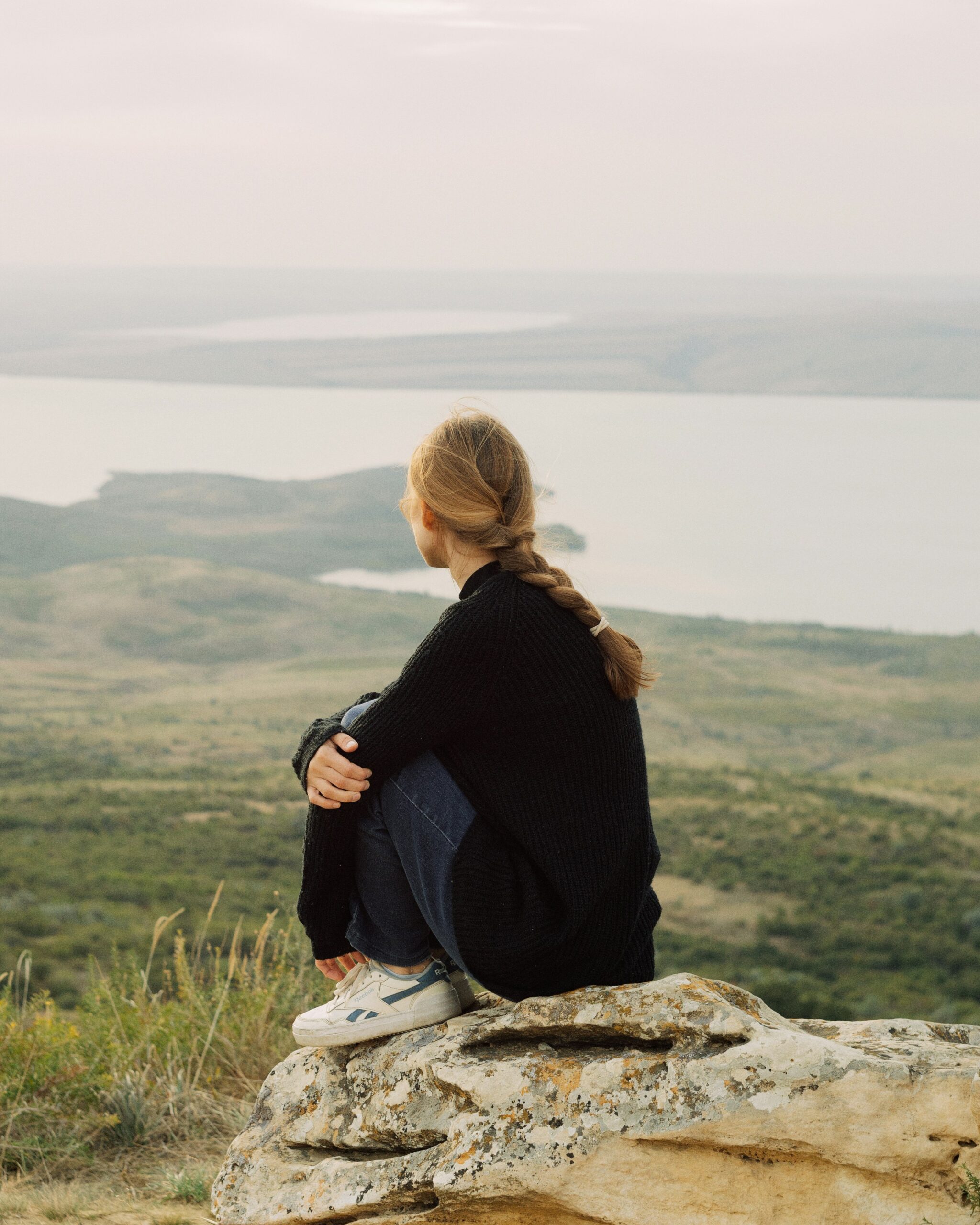Person with braided hair sitting alone on a rock above a still lake, weighing a decision about a relationship
