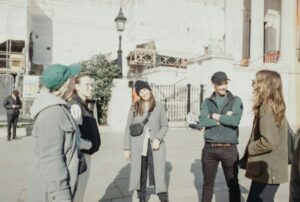 Five young adults in coats and beanies standing in a loose circle, talking and laughing on a sunny city sidewalk, the kind of small Christian community where everyone knows everyone