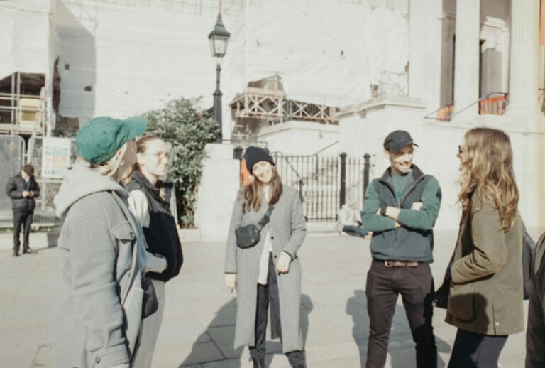 Five young adults in coats and beanies standing in a loose circle, talking and laughing on a sunny city sidewalk, the kind of small Christian community where everyone knows everyone