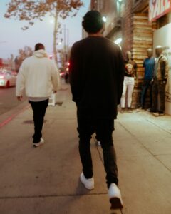 Two young men walking the same direction down a city sidewalk at dusk, one in a white hoodie a few steps ahead of the other in black, the pace and space to keep before dating a friend's ex