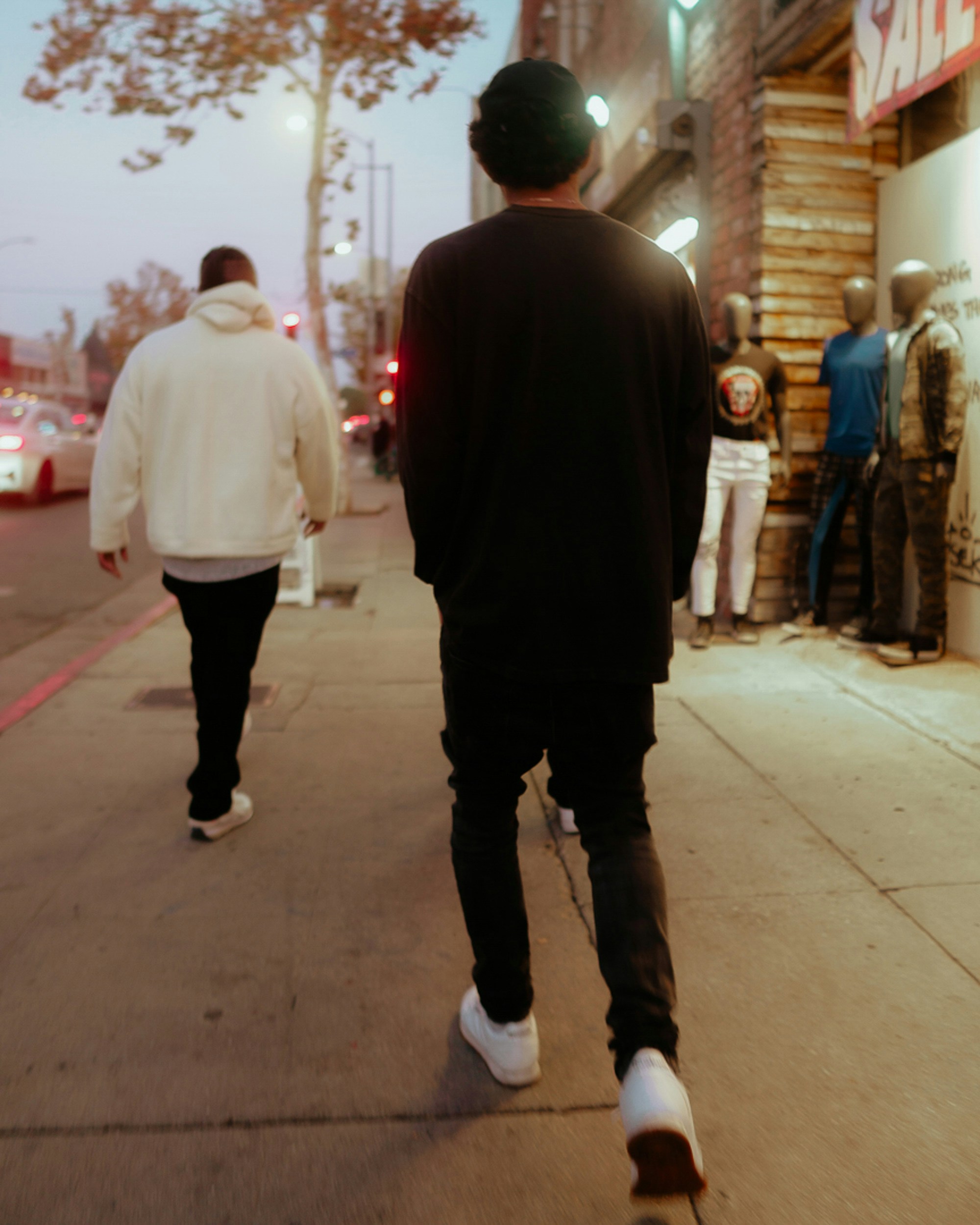 Two young men walking the same direction down a city sidewalk at dusk, one in a white hoodie a few steps ahead of the other in black, the pace and space to keep before dating a friend's ex