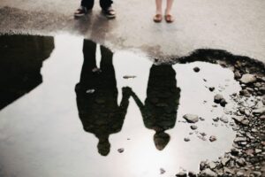 Reflection of a couple holding hands in a rain puddle on grey pavement, only their feet visible above it, a picture of being fully seen without shame in marriage