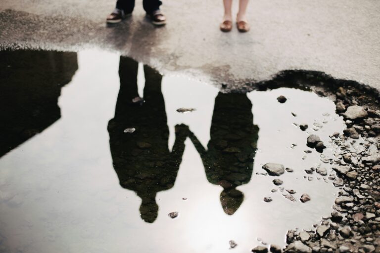 Reflection of a couple holding hands in a rain puddle on grey pavement, only their feet visible above it, a picture of being fully seen without shame in marriage