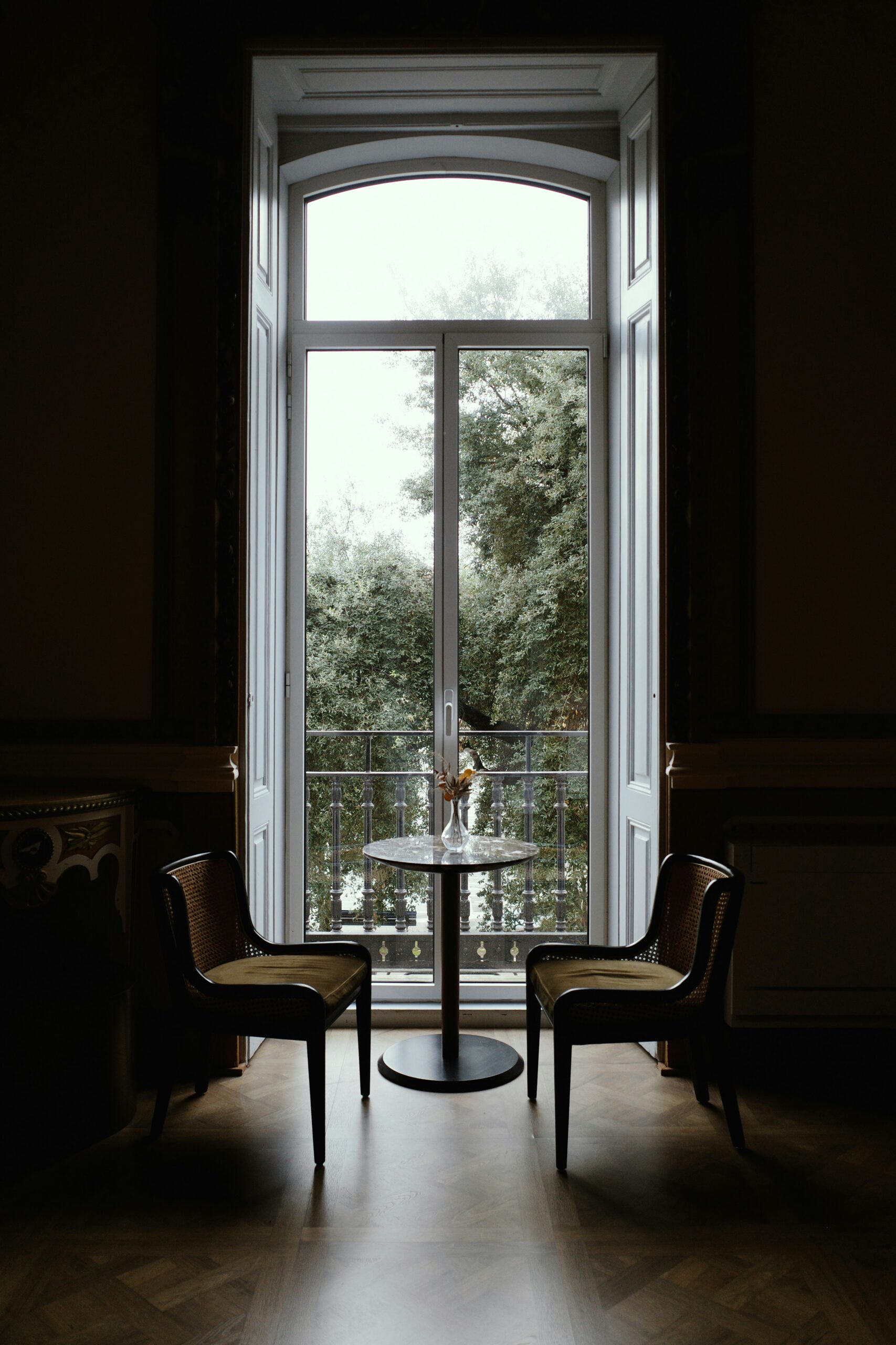 Two chairs and a small round table beside a tall window overlooking trees.