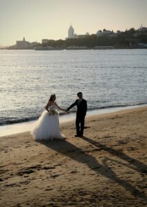 Bride and groom holding hands on a sandy beach beside the water.