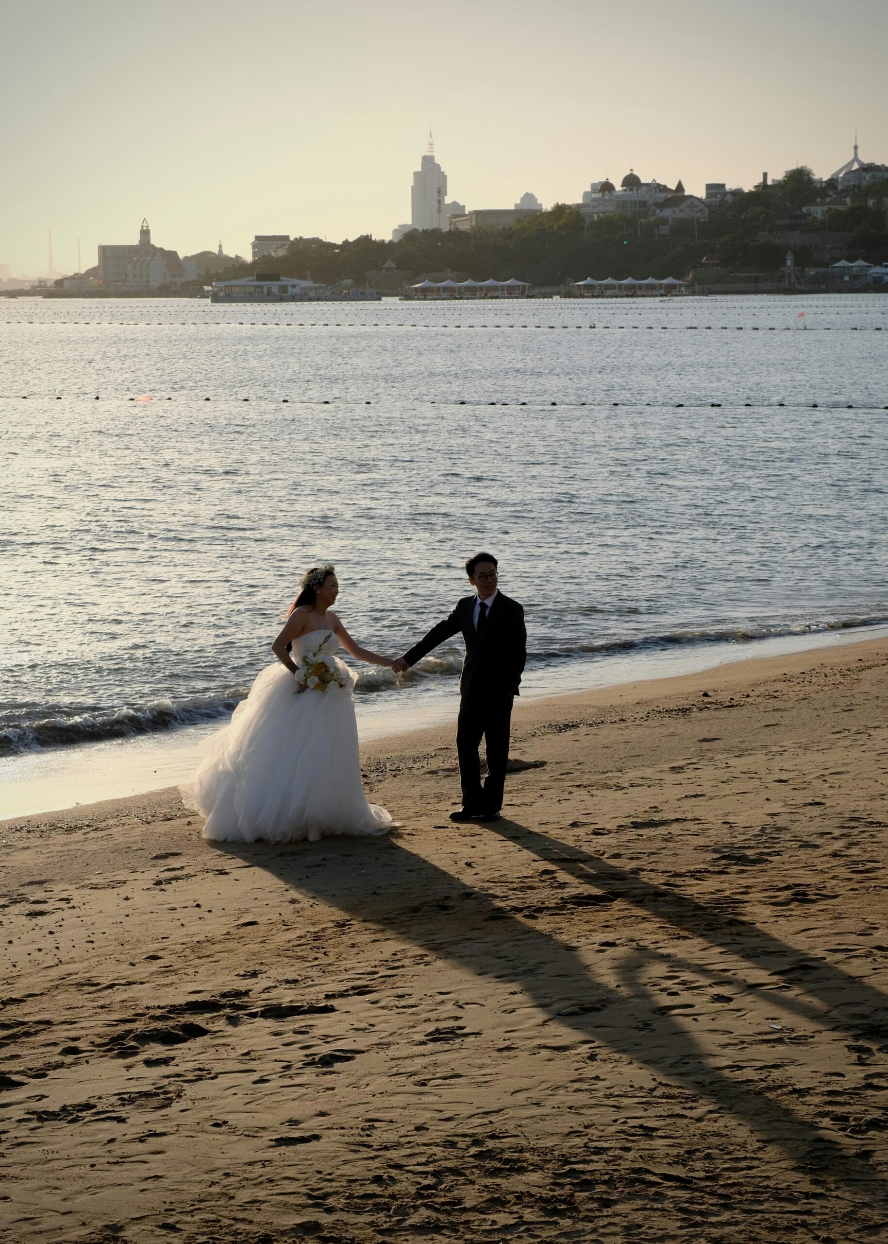 Bride and groom holding hands on a sandy beach beside the water.